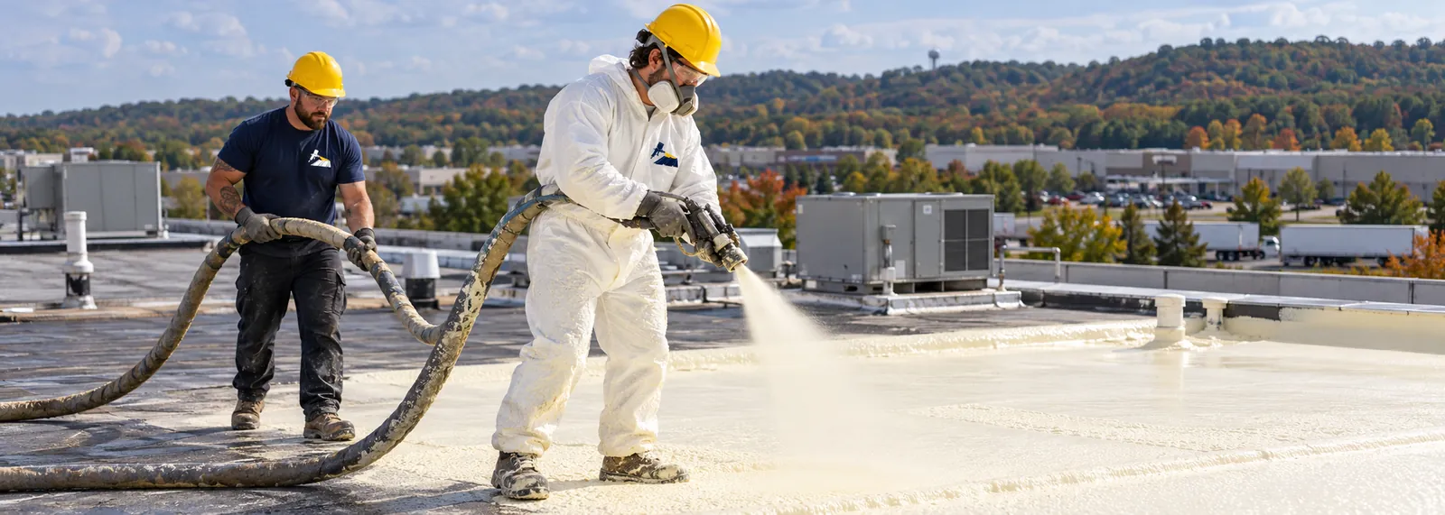 Sprayer in a protective suit applying foam over a commercial roof while a second worker feeds the hose